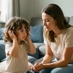 Une fillette de quatre ans se couvre doucement les oreilles tandis qu’une femme lui apporte du réconfort dans un salon calme et adapté aux sensibilités sensorielles.